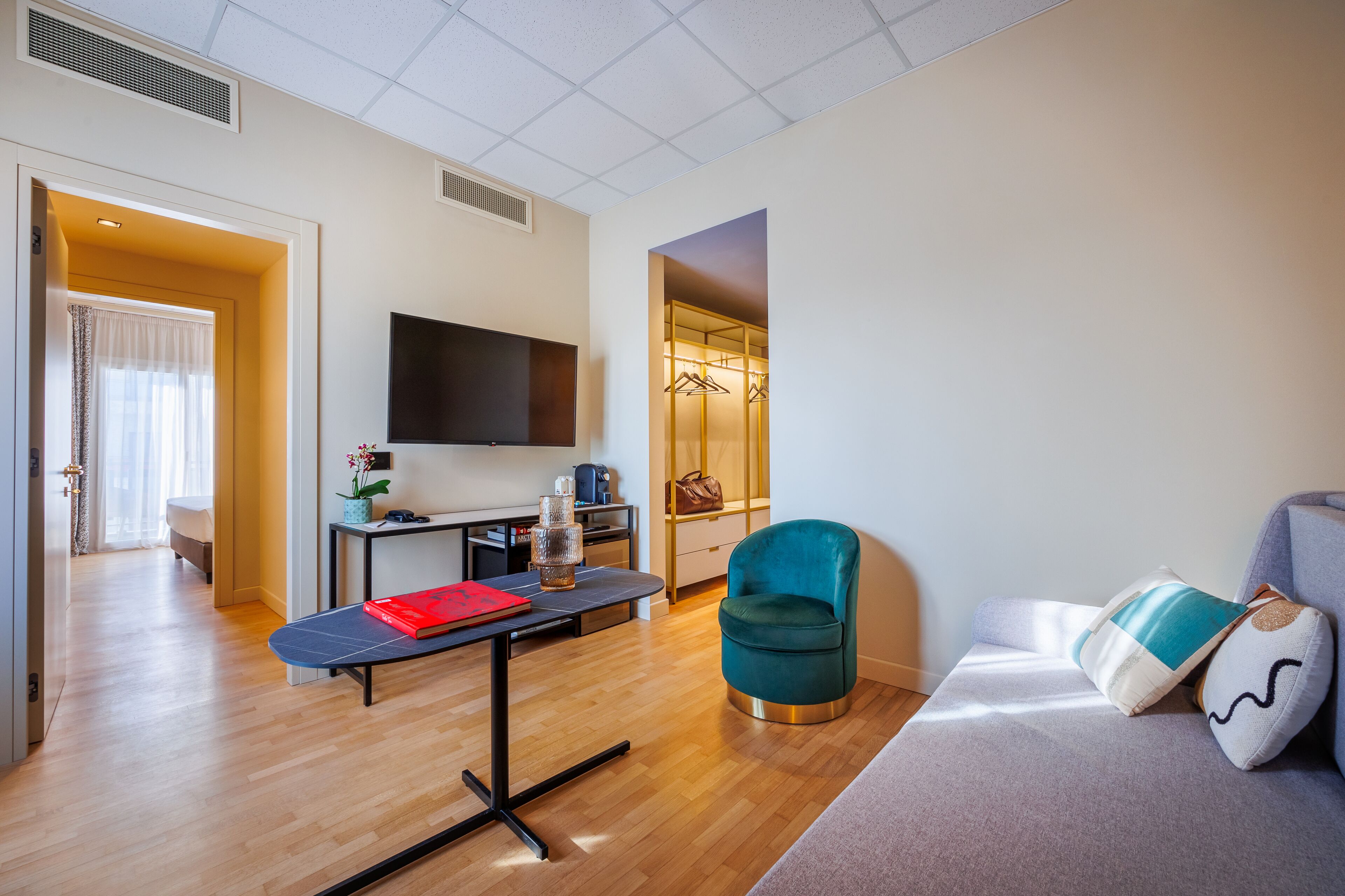 A modern hotel room with a wooden floor, a flat-screen TV mounted on the wall, a desk with a chair, a sofa with cushions, and a walk-in closet with hanging clothes.