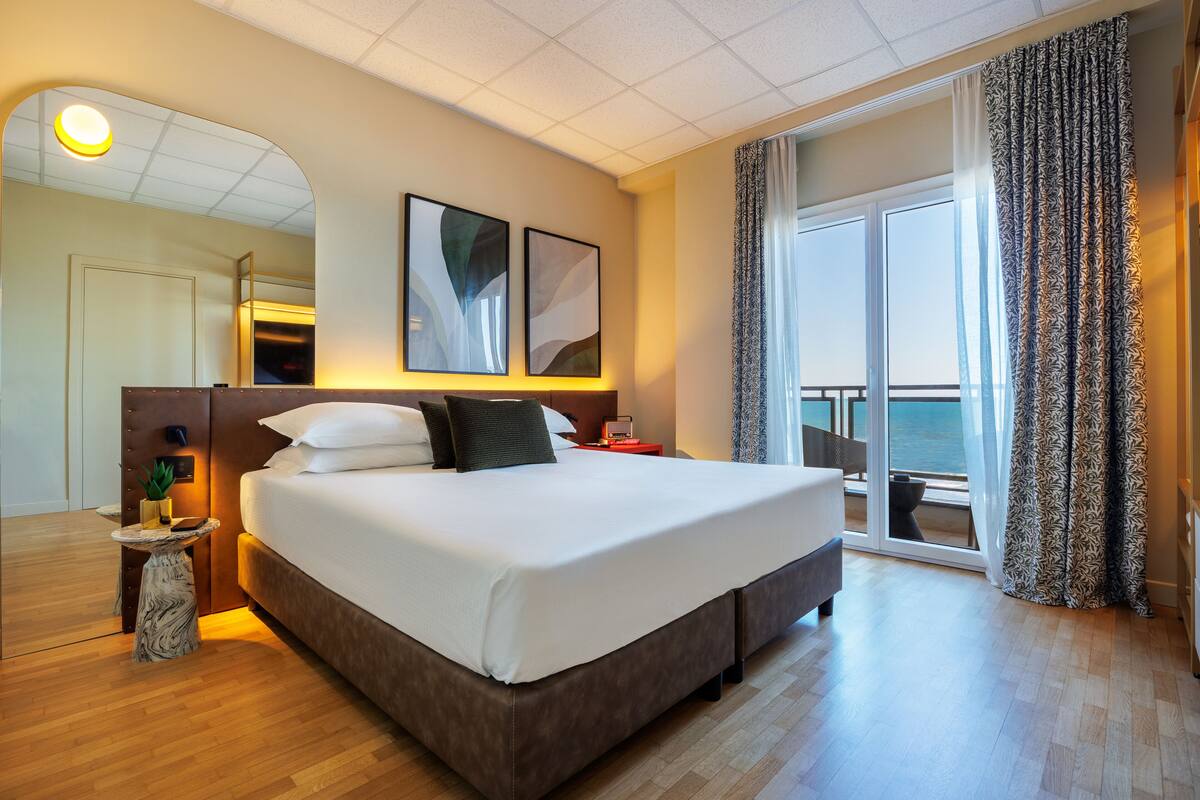 Habitación de hotel moderna con una cama grande, una mesita auxiliar, un espejo y vistas al mar desde el balcón.