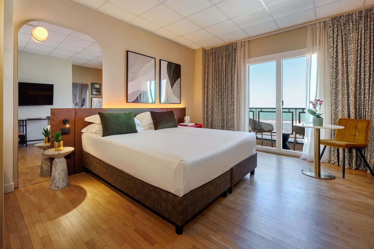 Habitación de hotel con una cama grande, una mesita con una planta, una silla y un balcón con vistas.