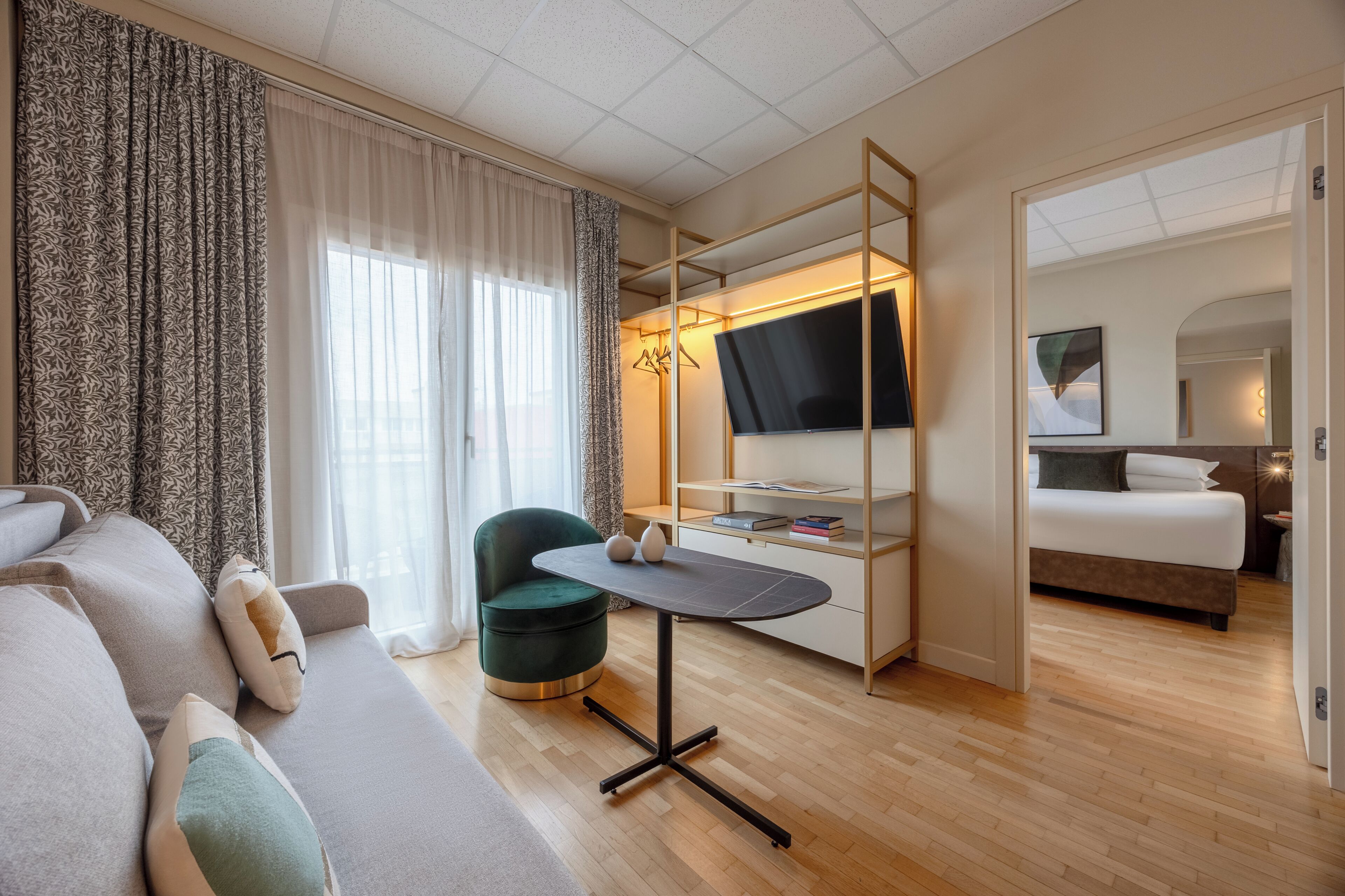 A modern hotel room with a sofa, a round table, a TV on a stand, a bed, and a wardrobe.