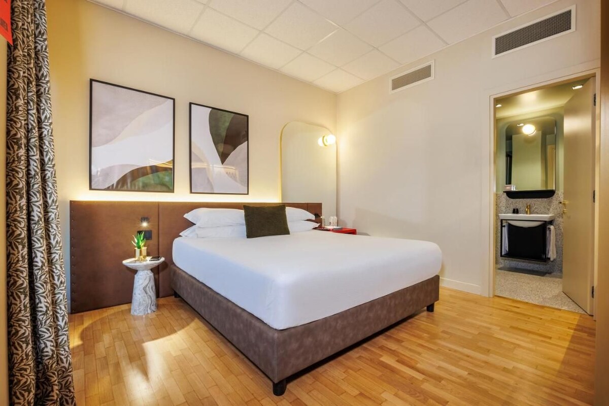 A hotel room with a large bed, bedside table, lamp, and artwork on the wall.