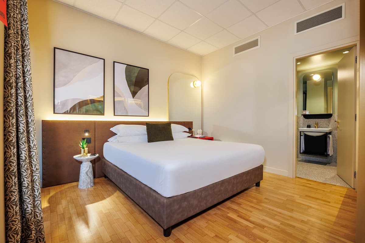 Una habitación de hotel con una cama grande, una mesita de noche con lámpara, una pequeña mesita con una planta, y un baño visible a través de una puerta abierta.