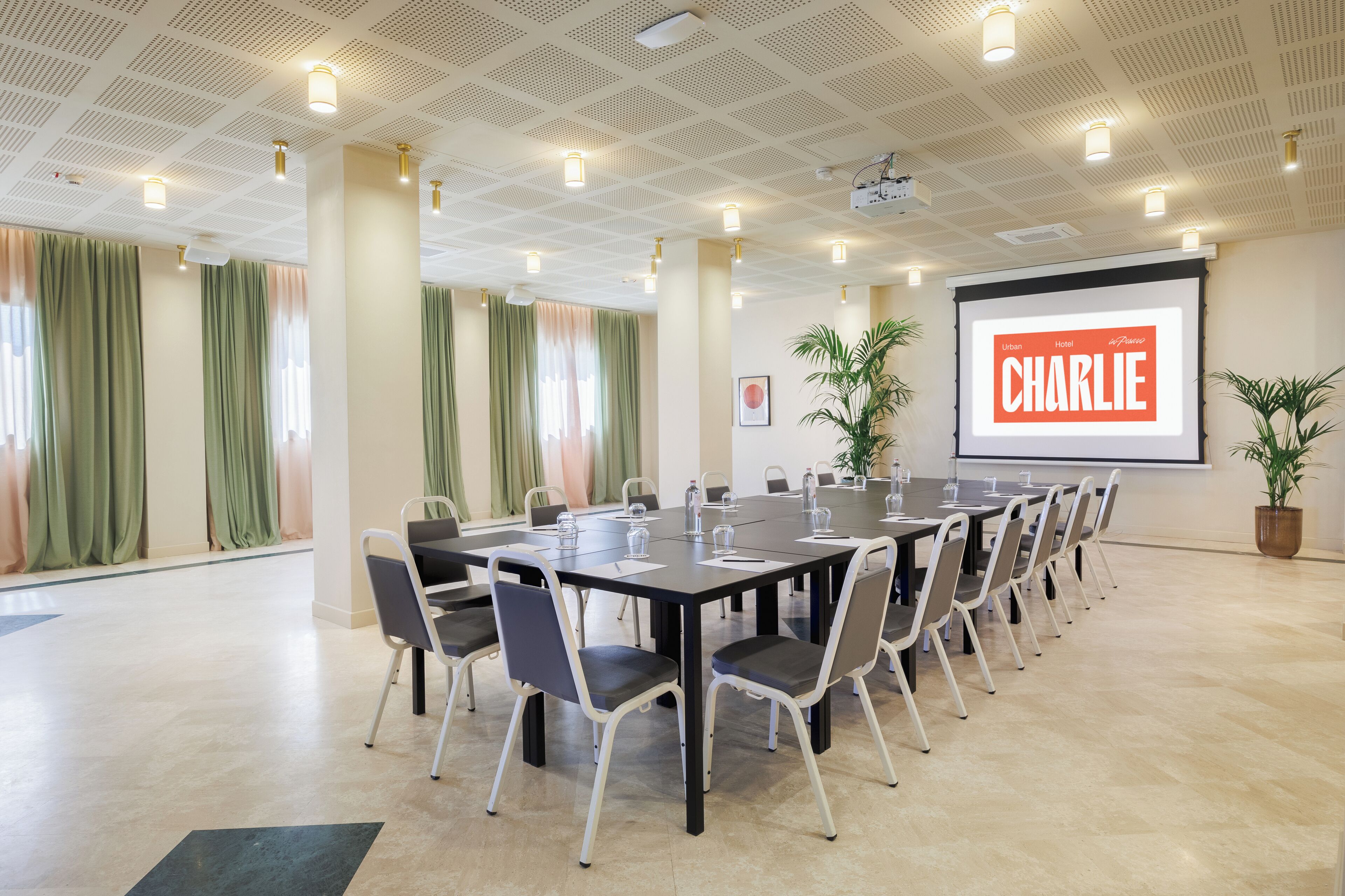 Una sala conferenze con un lungo tavolo, sedie, uno schermo per proiettore che mostra "CHARLIE" e piante in vaso.