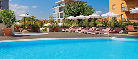 A pool area with lounge chairs and umbrellas, surrounded by buildings and greenery.