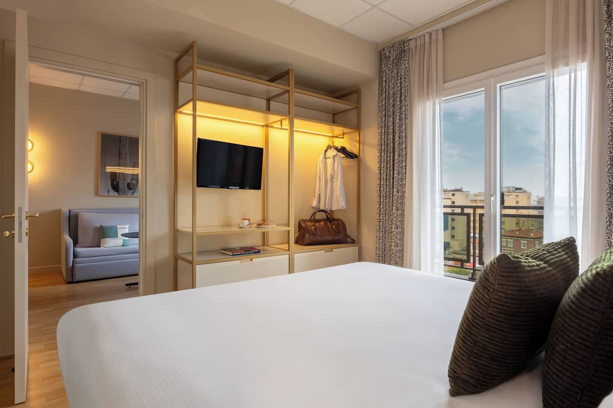 Habitación de hotel con una cama grande, un vestidor con televisor y vistas a edificios a través del ventanal.