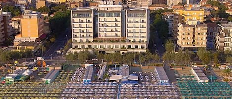 An aerial view of a coastal city with a large hotel featuring multiple floors and balconies, a beach with rows of umbrellas, and a parking lot.