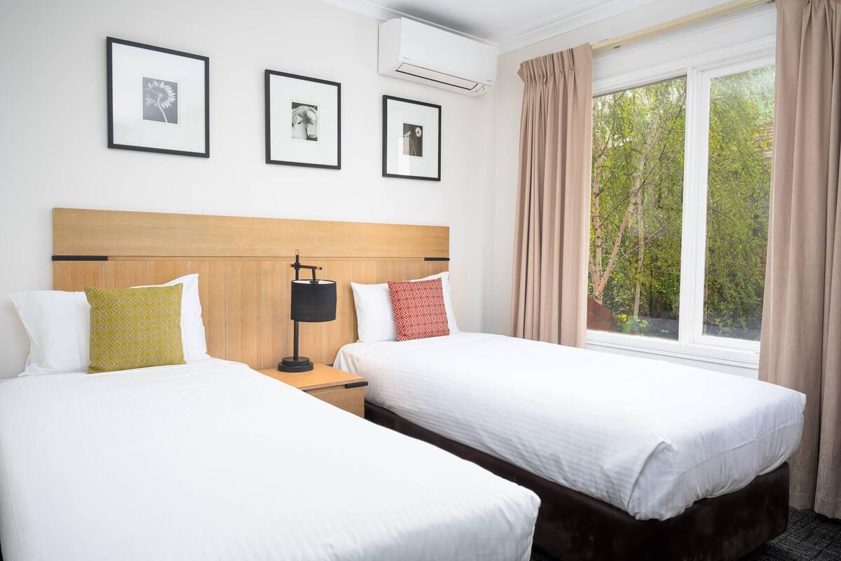 Habitación de hotel con dos camas, cabecera de madera, mesita de noche con lámpara y cuadros en la pared.