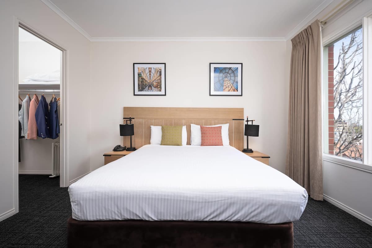 Una habitación de hotel con una cama grande, dos mesitas de noche con lámparas, un armario y dos cuadros en la pared.