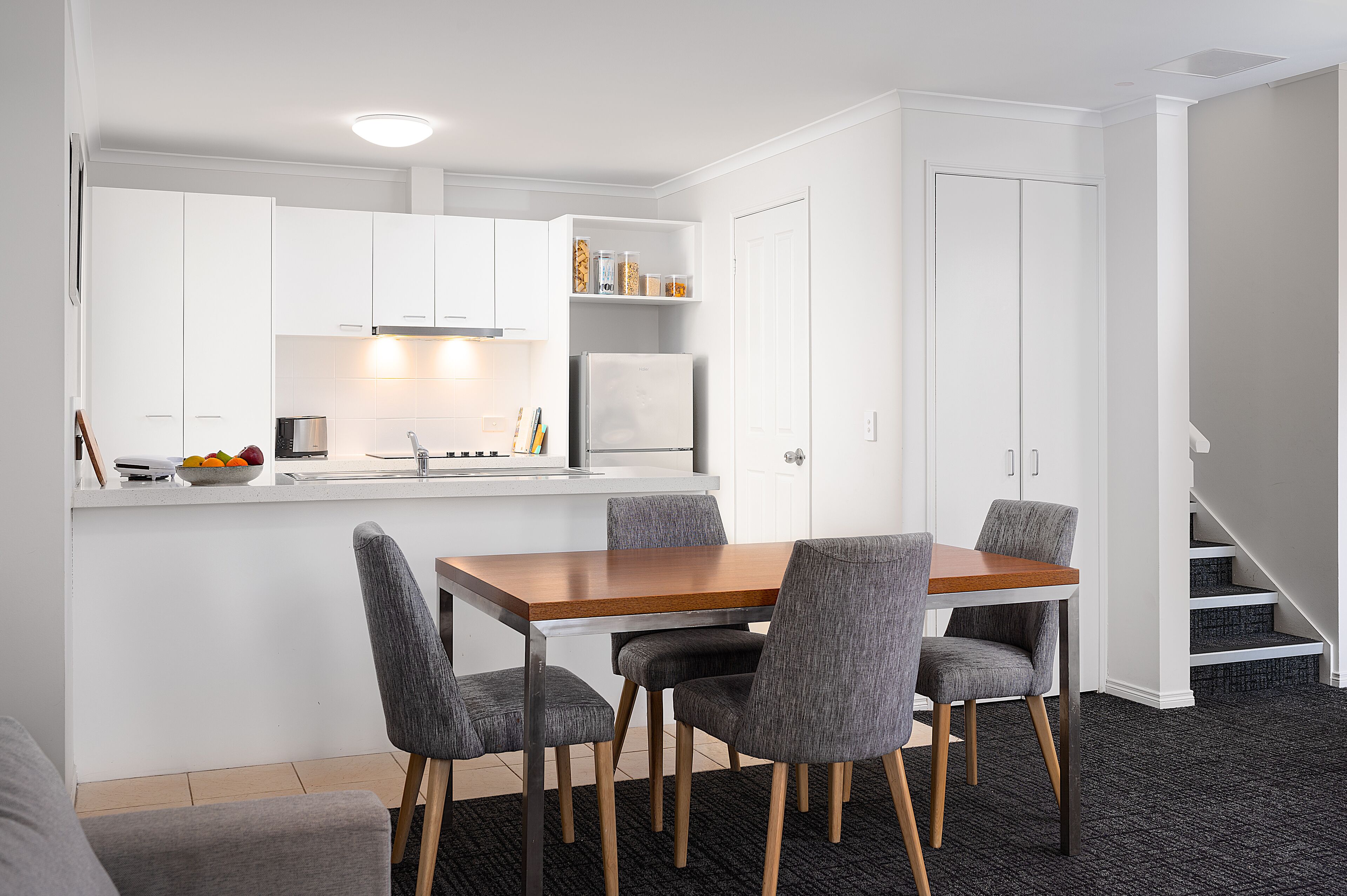 A compact kitchen with white cabinets, a wooden dining table, and four grey chairs.