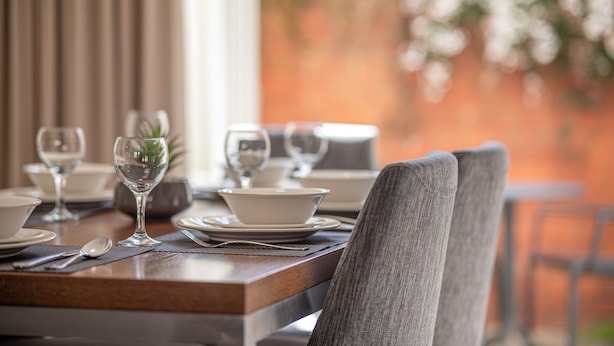 Mesa de jantar posta com pratos, tigelas e talheres brancos, com uma planta em um vaso de vidro.