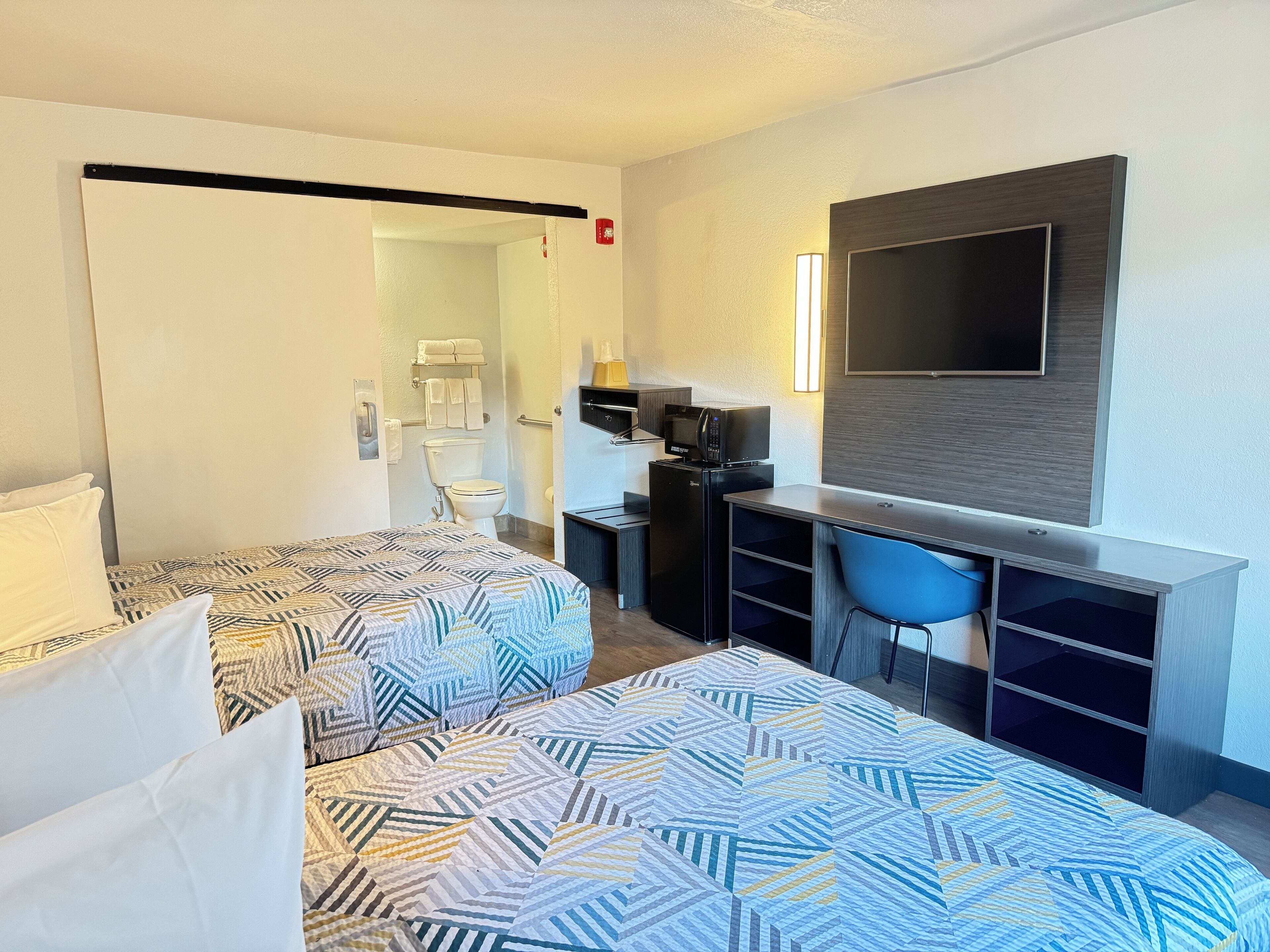 A hotel room with two beds, a desk with a chair, a TV, and a small kitchenette.