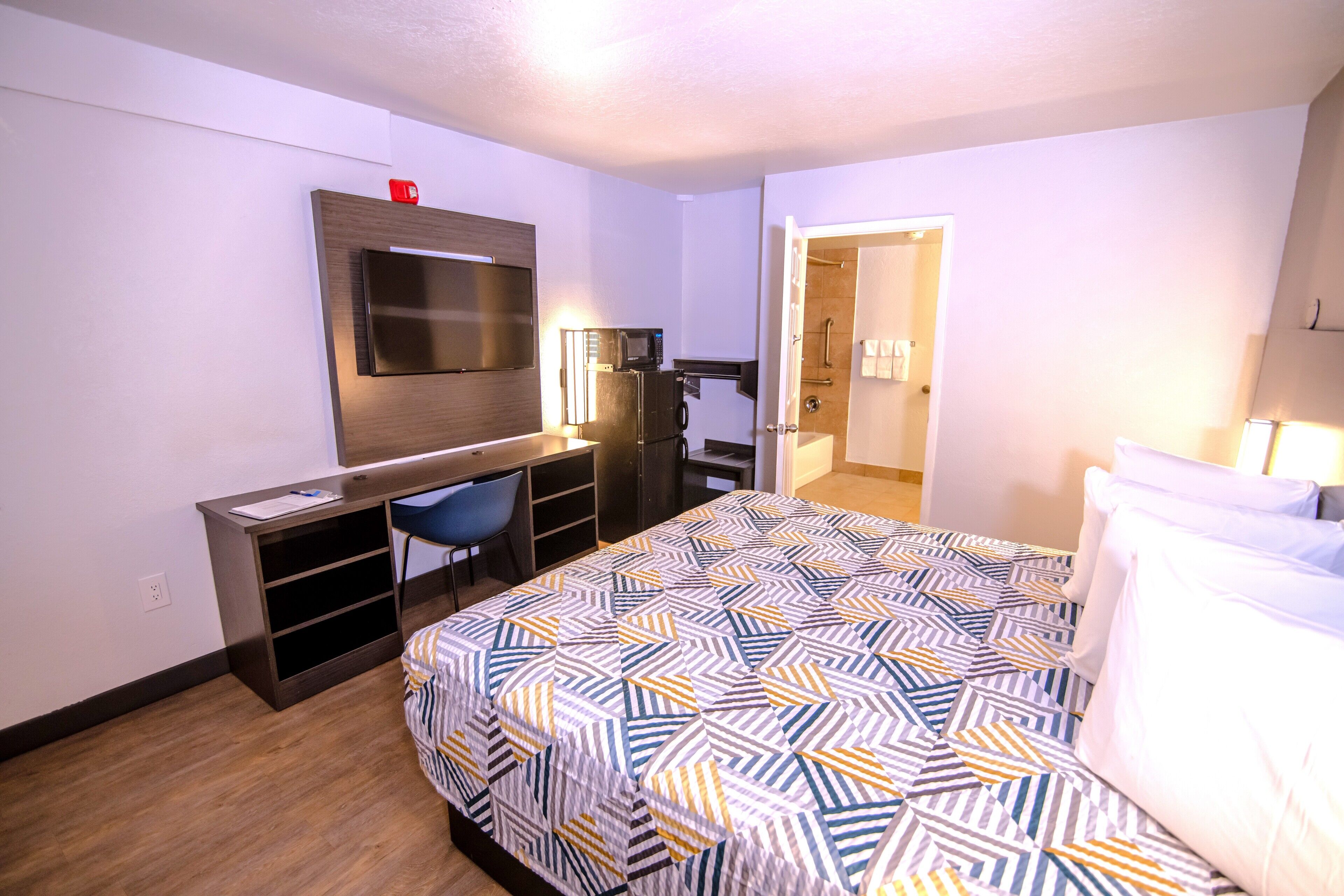 A hotel room with a bed, a desk, a chair, a TV, and a bathroom visible through an open door.