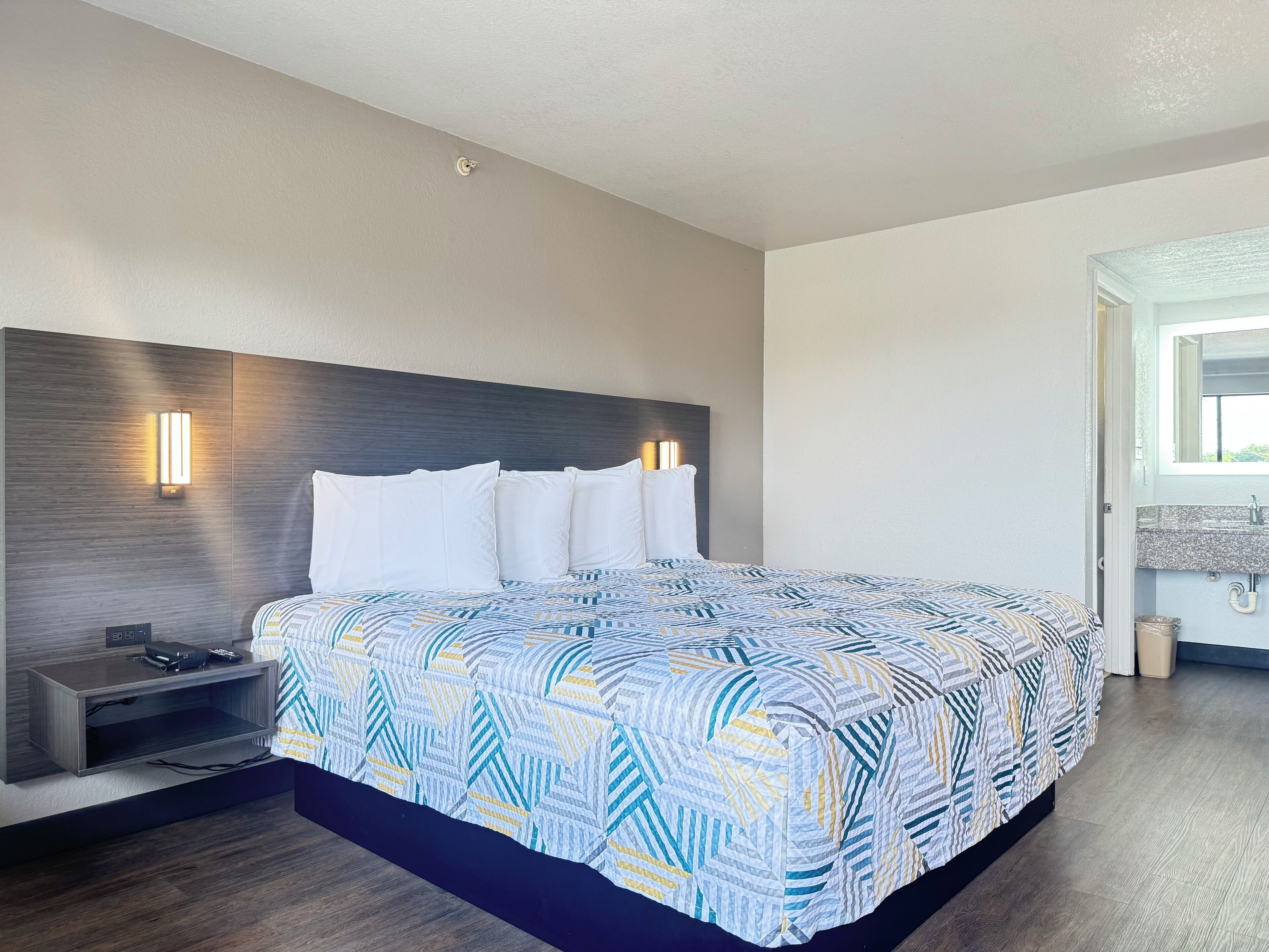 A modern hotel room with a large bed, a nightstand, a wall-mounted TV, and a bathroom visible through an open door.