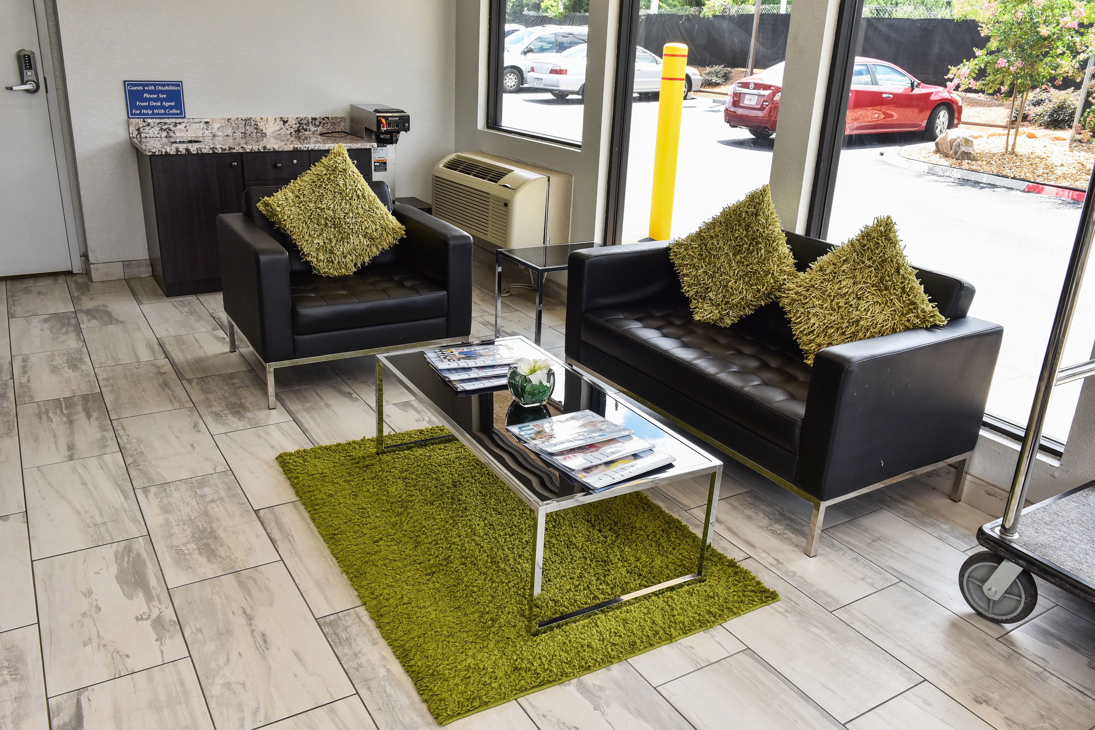 A modern waiting area with two black leather sofas, a green rug, a coffee machine, and a window with a view of parked cars.