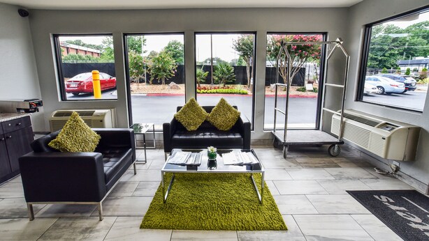 A modern hotel lobby with a black sofa, a glass coffee table, and a green rug. There are large windows offering a view of the parking lot and greenery outside. - Masters Inn Tucker (Decatur)