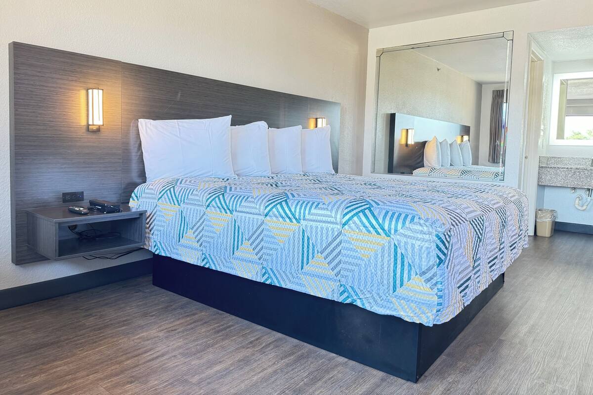 Una habitación de hotel moderna con una cama grande, una mesita de noche con lámpara y un espejo sobre la cama.