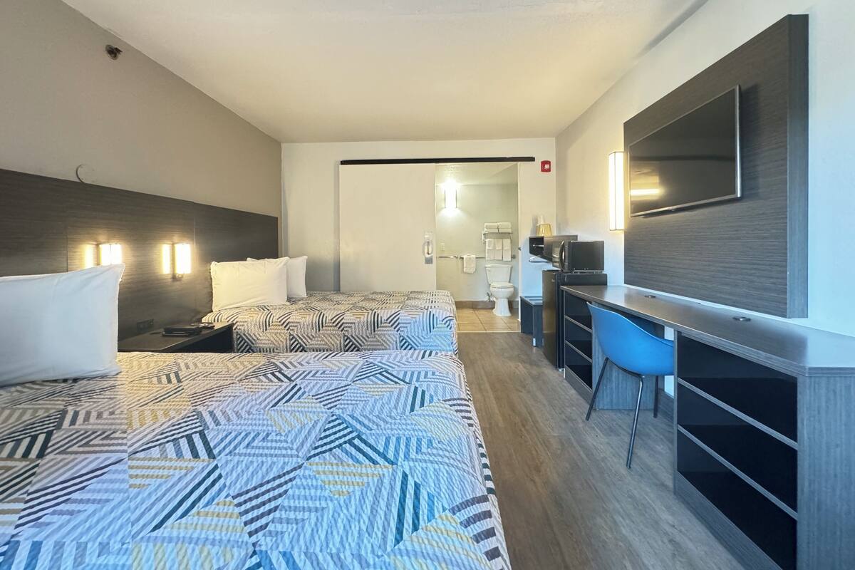Habitación de hotel con dos camas, un escritorio, una silla, un televisor y un baño visible a través de una puerta abierta.