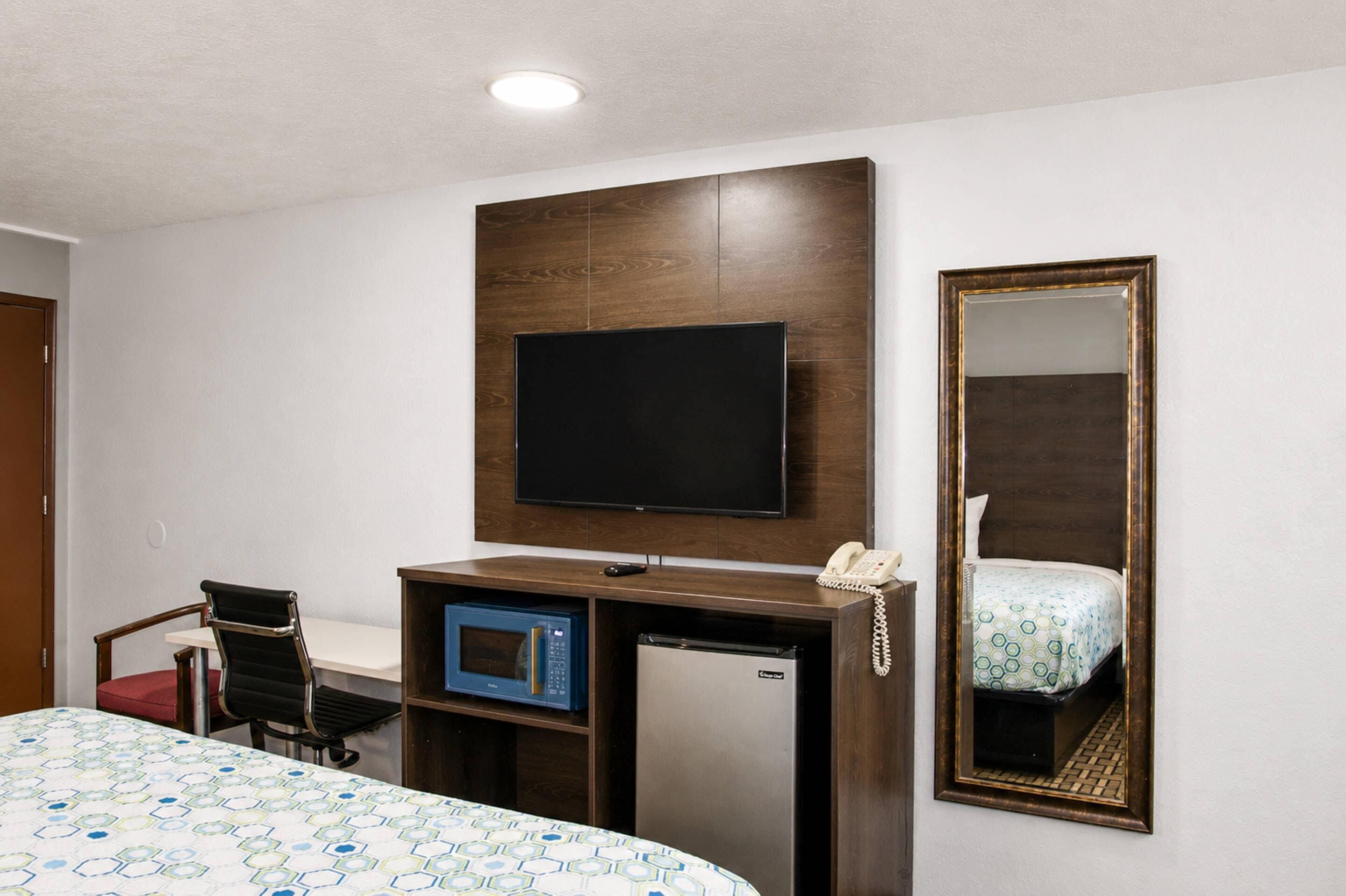A hotel room with a bed, a desk with a computer and a telephone, a microwave, a television, and a large mirror.