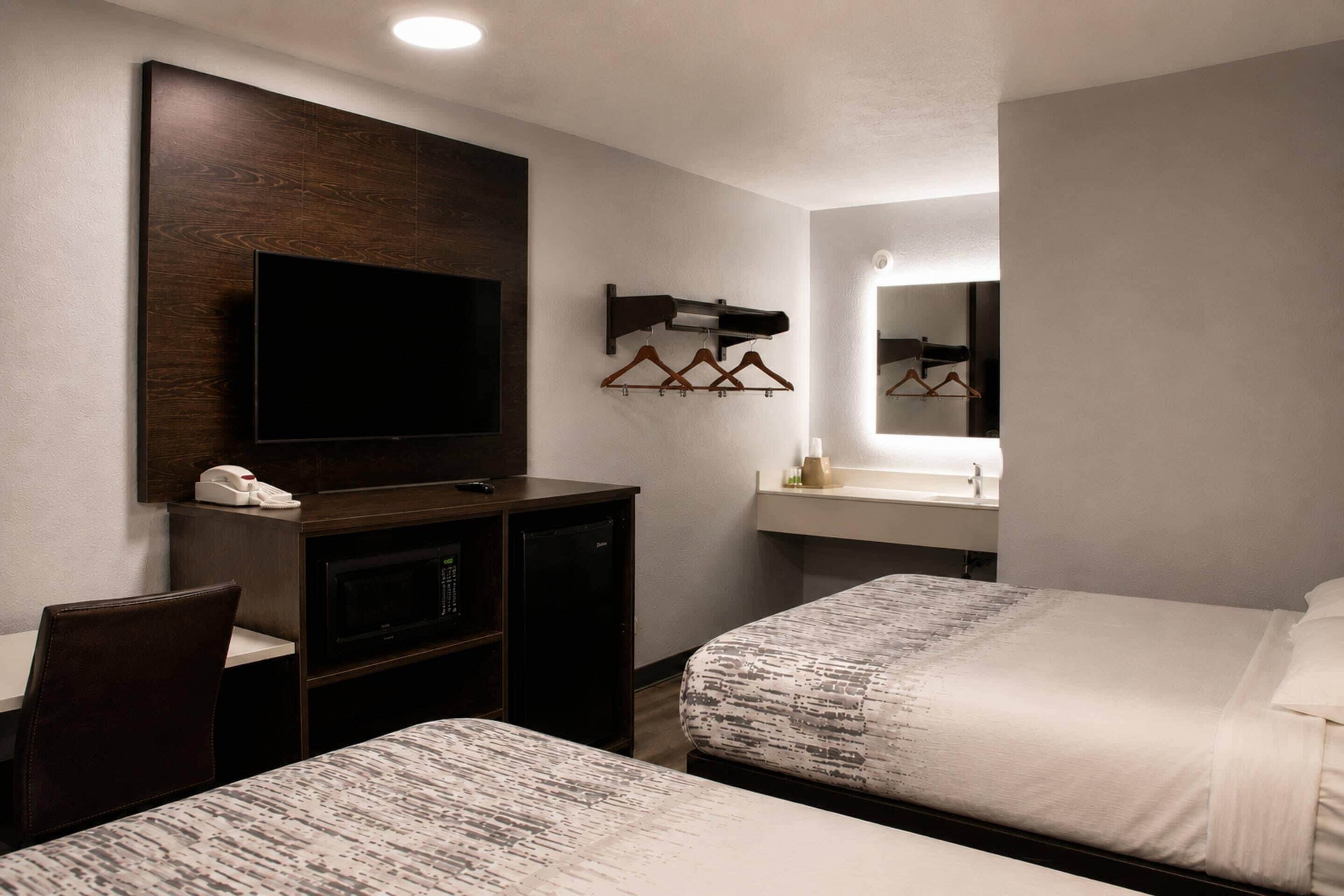 Habitación de hotel con dos camas, un escritorio con televisor, un espejo y un baño pequeño.