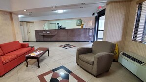 Una hall d'albergo con un divano rosso, una poltrona marrone, un tavolino da caffè, un banco reception e un pavimento decorato.