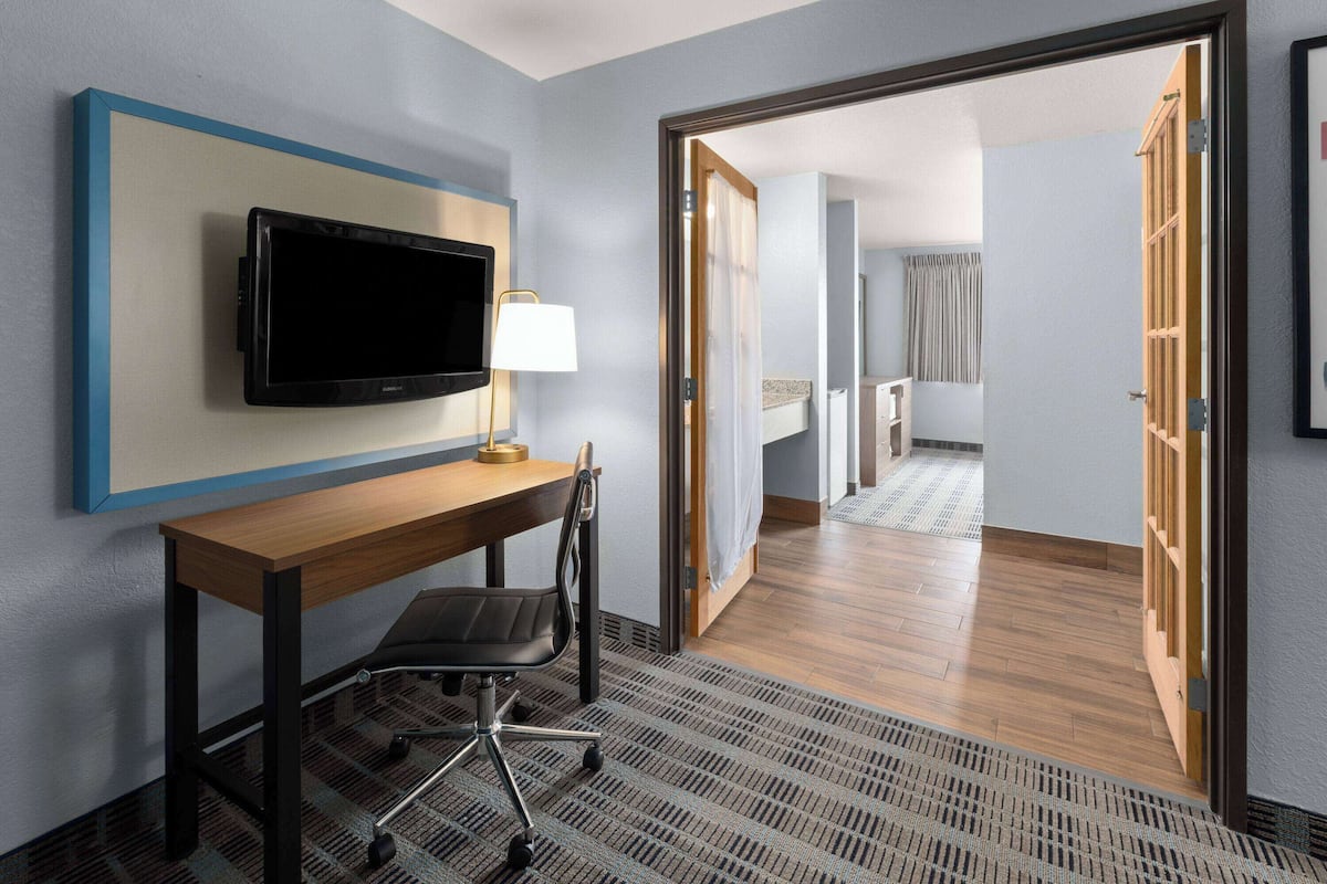 Ein Hotelzimmer mit Holztisch, schwarzem Bürostuhl, fest installiertem Flachbildfernseher und einer Lampe.