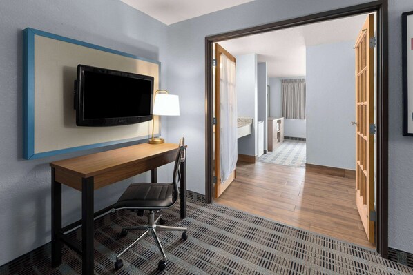 A hotel room with a wooden desk, a black office chair, a mounted flat-screen TV, and a lamp.