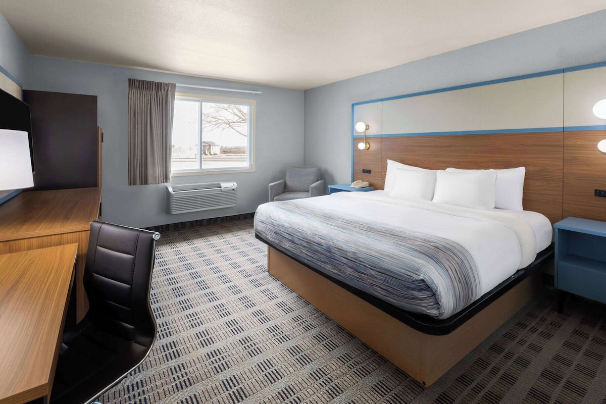 A hotel room with a large bed, a desk, a chair, a small table, a nightstand, and a window with curtains.