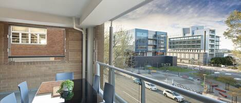 A balcony with a table and chairs, a view of a parking lot, and modern buildings in the background.
