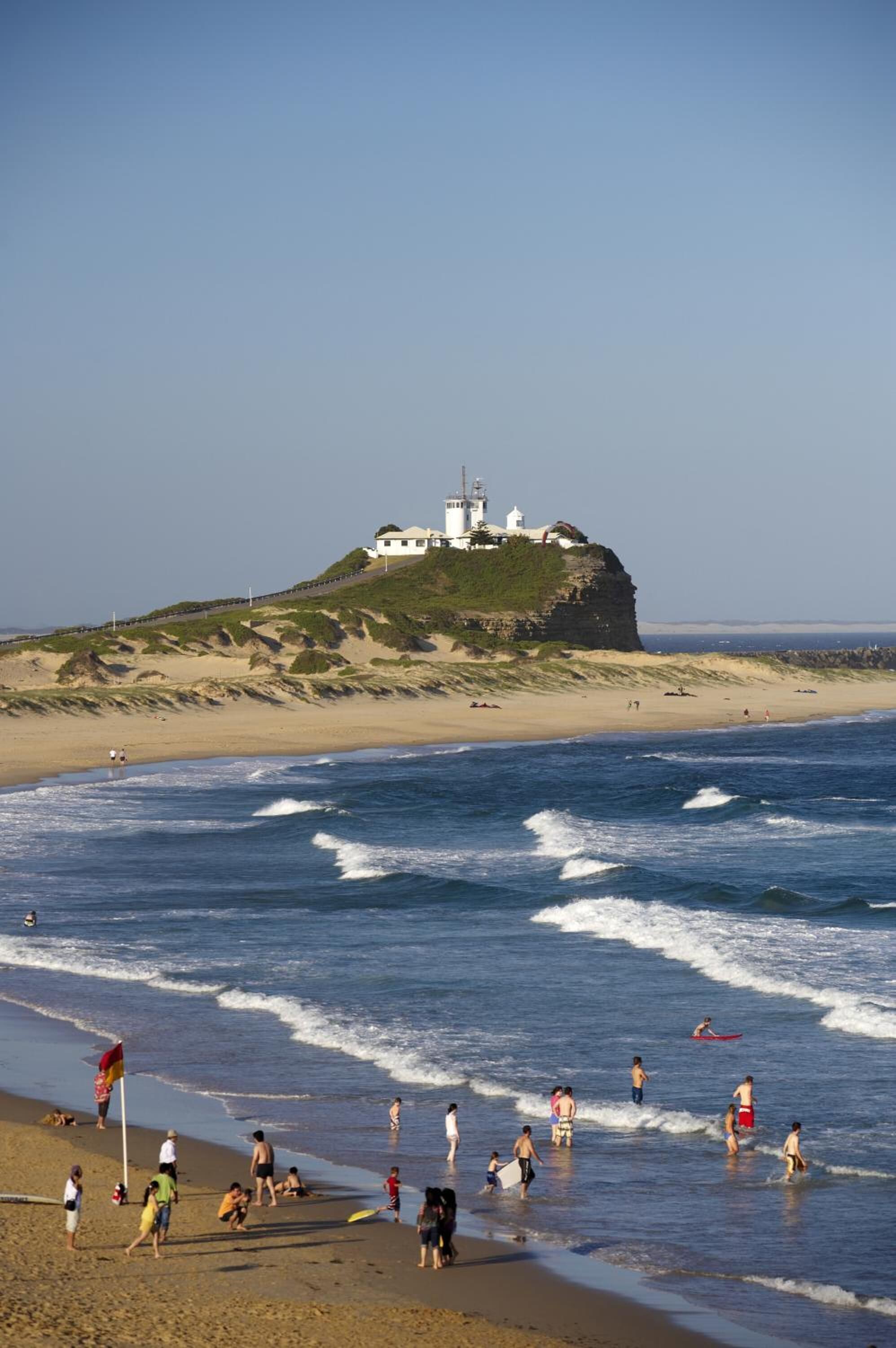 A beach with a lighthouse on a cliff, waves, and people enjoying the beach.
