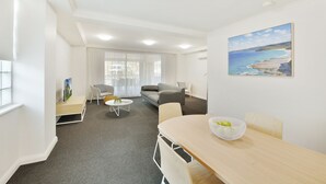 En moderne stue med sofa, sofabord og spisebord. Et billede hænger på væggen, og et vindue er dækket af gardiner.