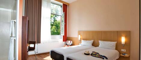 A hotel room with two beds, a window with curtains, a ceiling fan, and a bedside table with a lamp.