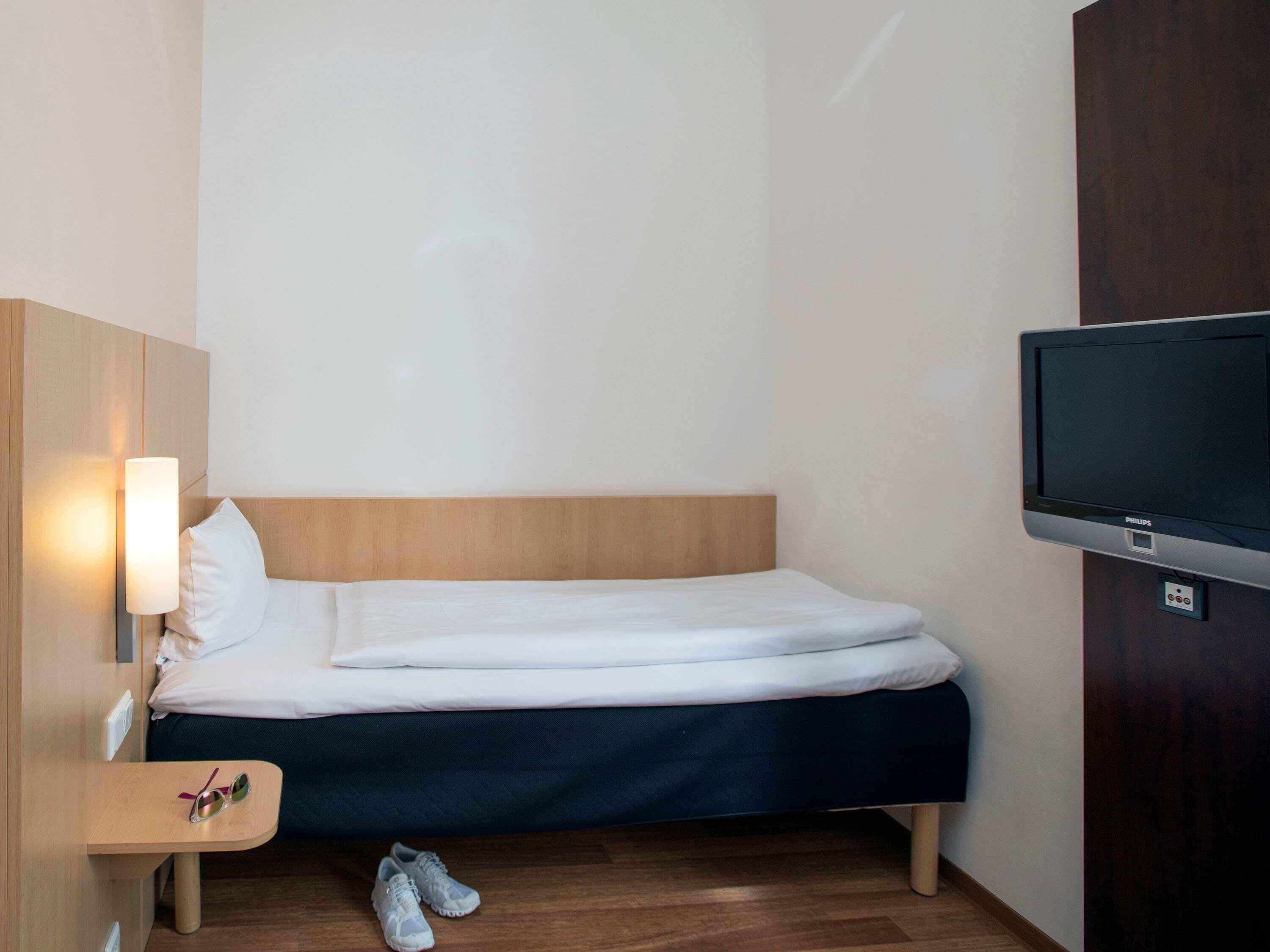 Een eenpersoonsbed met houten hoofdbord, een nachtkastje met lamp, een paar schoenen en een televisie aan de muur.