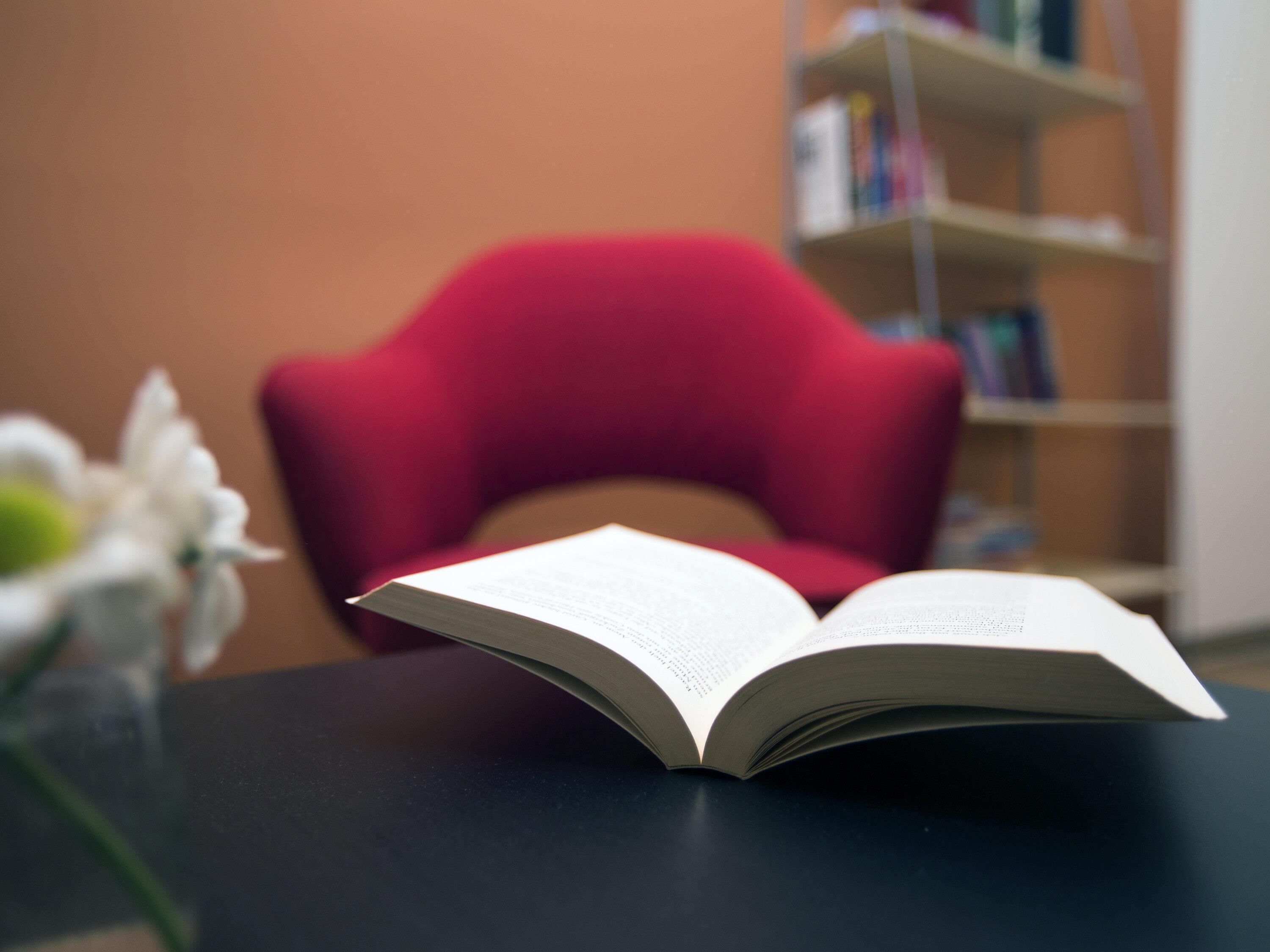 An open book on a dark surface with a red chair and a bookshelf in the background.