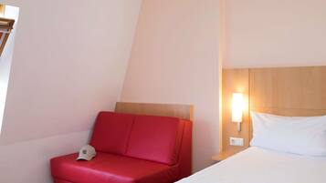 Una habitación de hotel con un sofá rojo, una cama con ropa de cama blanca y una lámpara fijada a la pared.