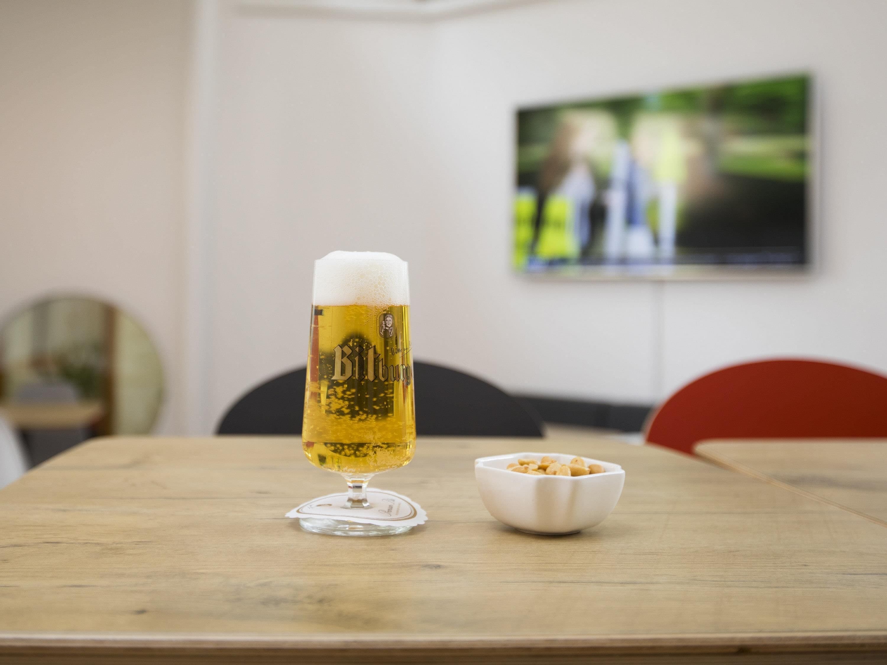 A beer glass on a wooden table with a bowl of peanuts beside it.