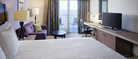 A hotel room with a large bed, a desk with a flat-screen TV, a chair, a small table, and a balcony with a view.