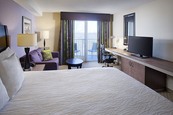 A hotel room with a large bed, a desk with a flat-screen TV, a chair, a small table, and a balcony with a view. - Hilton Garden Inn Hartford North/Bradley Int'l Airport (Windsor)