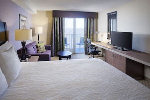 A hotel room with a large bed, a desk with a flat-screen TV, a chair, a small table, and a balcony with a view.