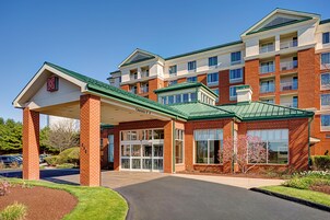 A multi-story hotel with a covered entrance, brick facade, and a parking area with cars.