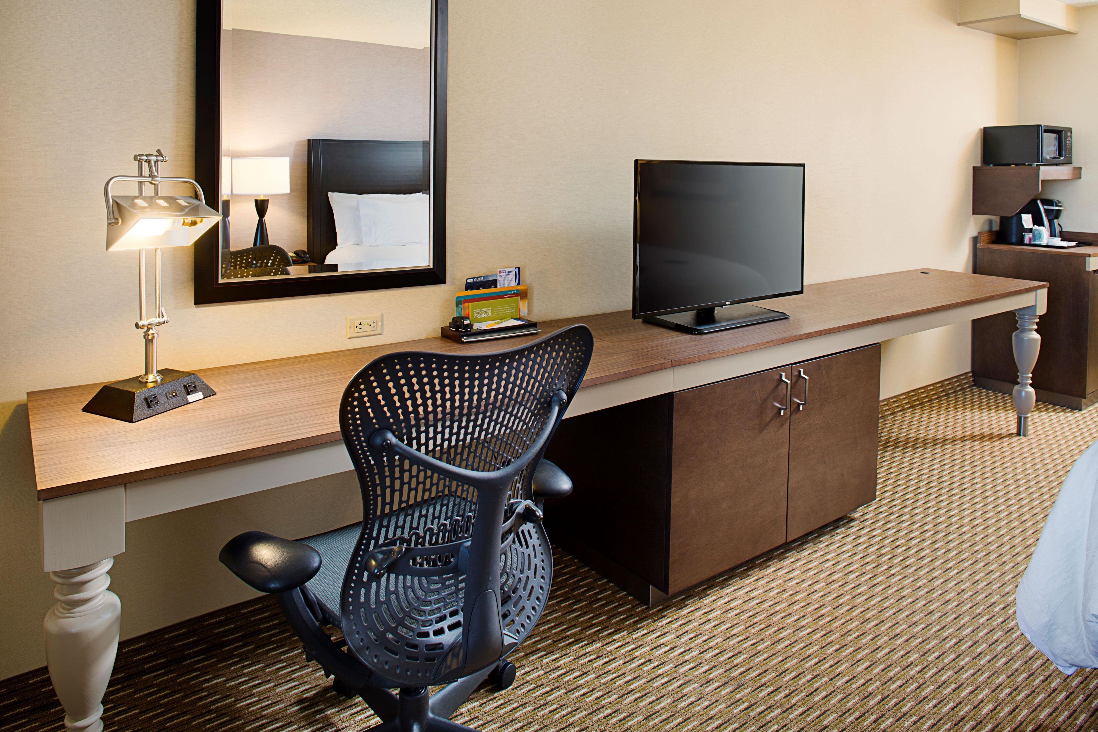 A hotel room with a desk, a chair, a television, a mirror, and a coffee machine.