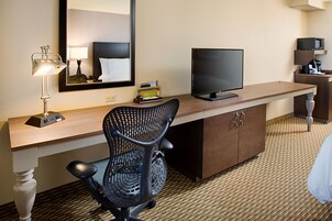 A hotel room with a desk, a chair, a television, a mirror, and a coffee machine.