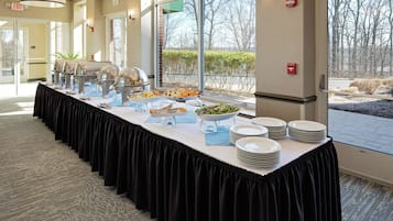Mesa de buffet con diversos alimentos y platos, un mantel negro y vistas a árboles a través de amplias ventanas.