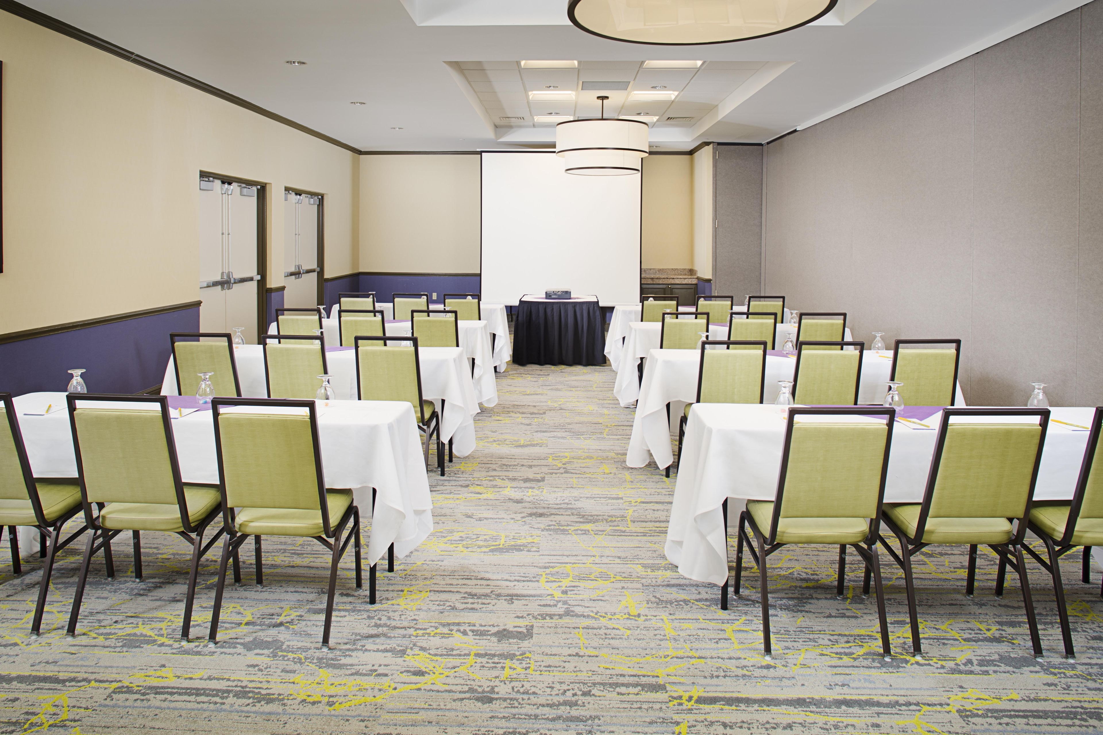Une salle de conférence avec des rangées de tables et de chaises, une scène à l’avant et un sol recouvert de moquette.