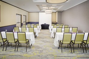 A conference room with rows of tables and chairs, a stage at the front, and a carpeted floor. - Hilton Garden Inn Hartford North/Bradley Int'l Airport (Windsor)