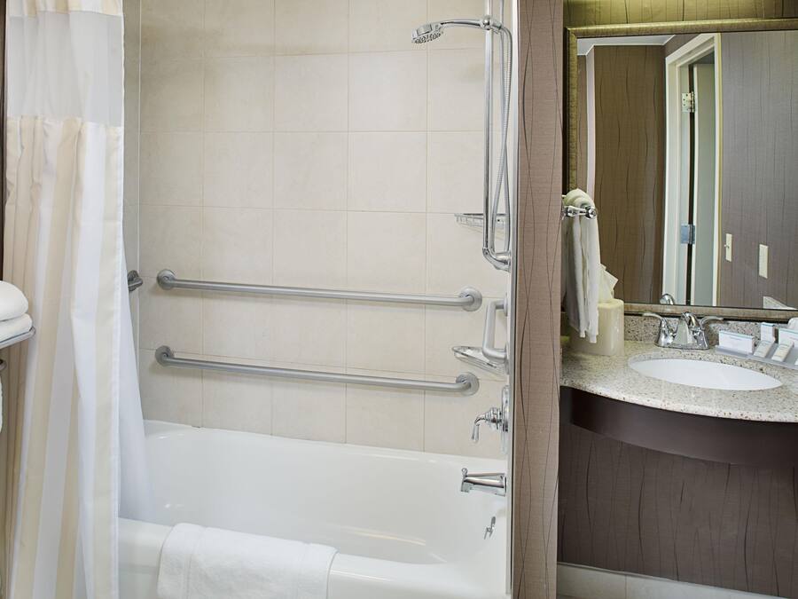 Un baño con bañera, ducha y lavamanos con espejo. Cuenta con barras de apoyo para mayor seguridad.