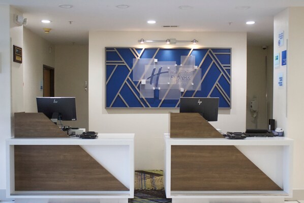 Una mesa de recepción con dos monitores de computadora, un adorno mural geométrico azul y un letrero de Holiday Inn Express & Suites.