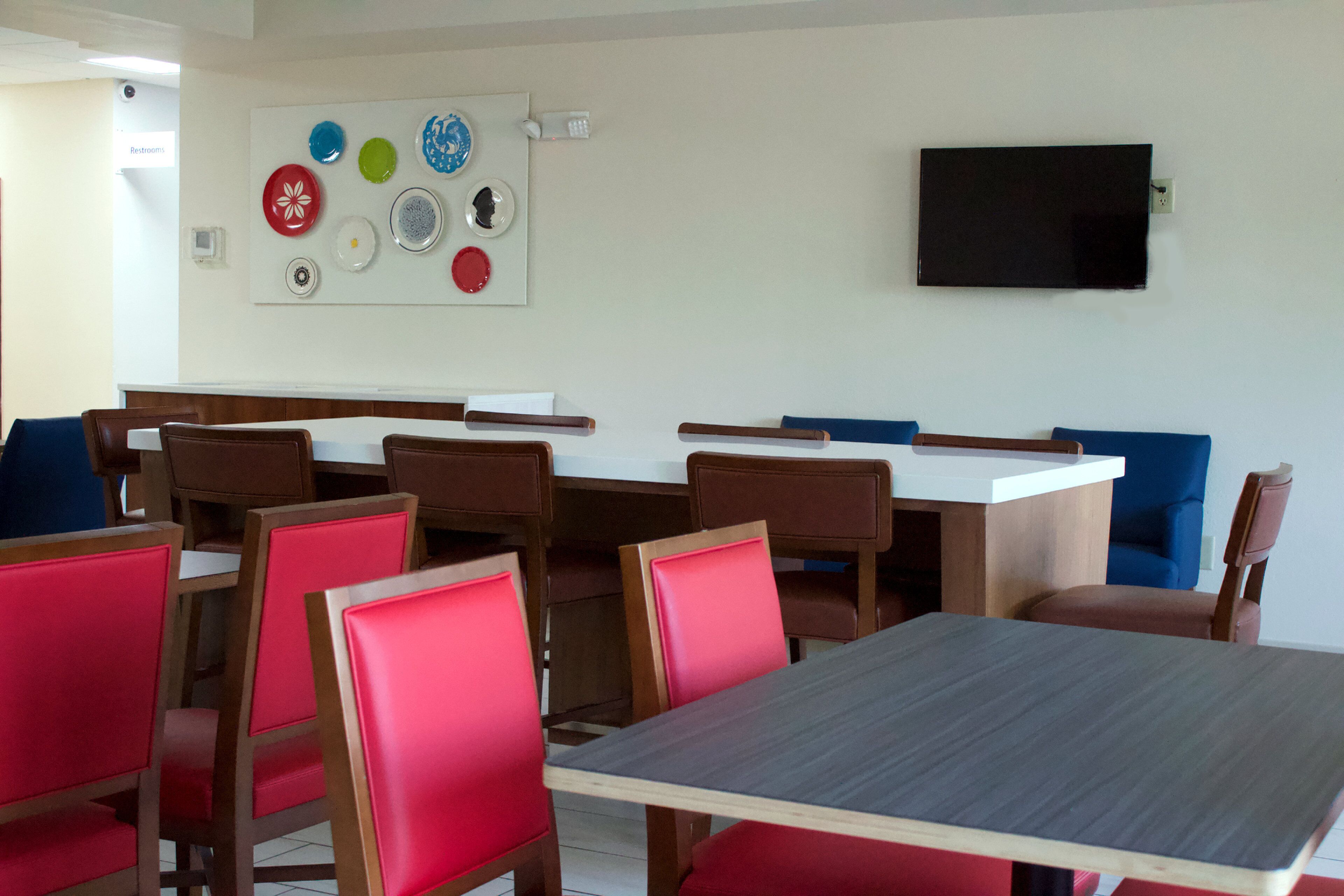 A dining area with wooden tables and red chairs, a whiteboard with colorful plates, and a flat-screen TV on the wall.