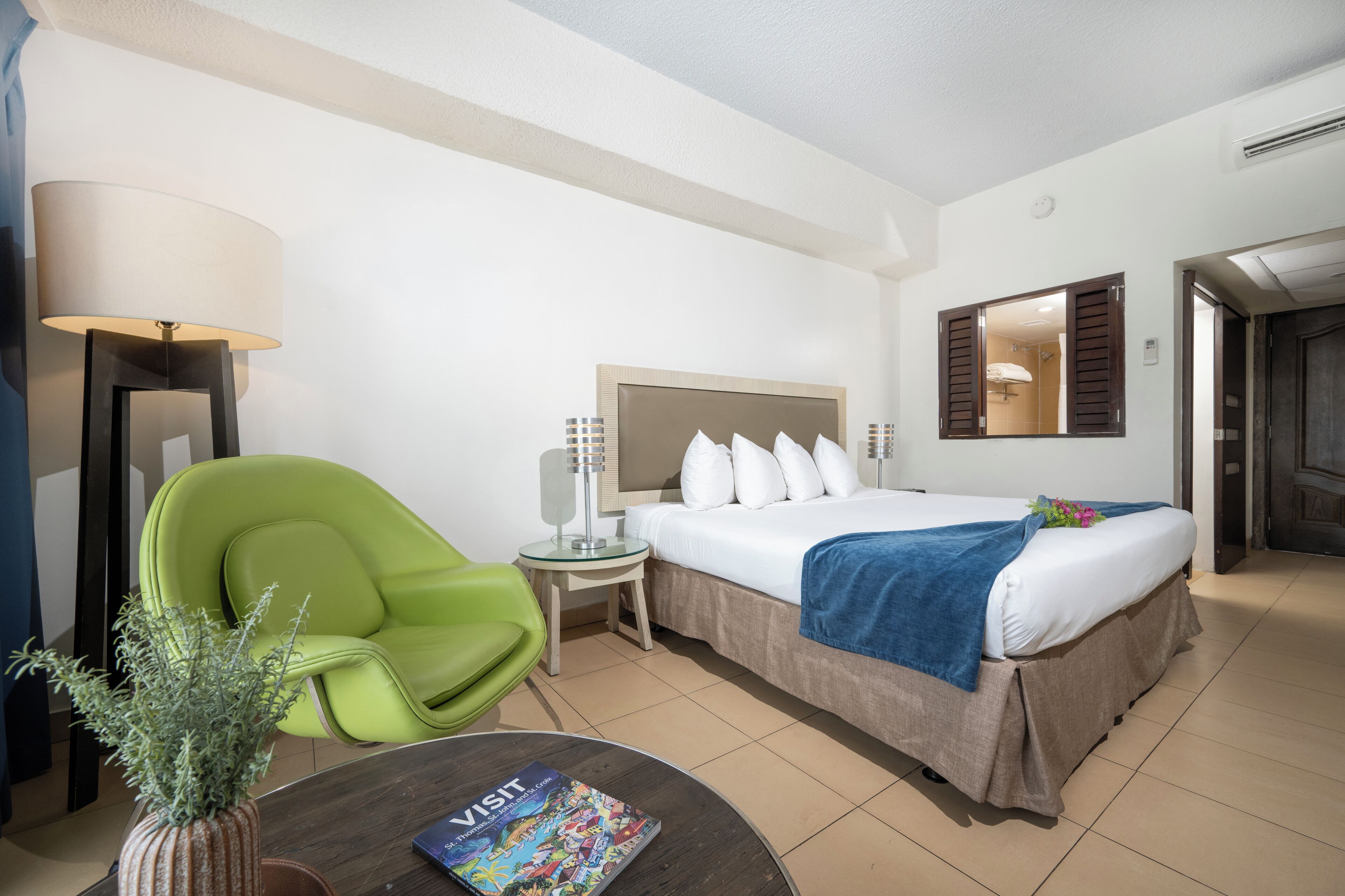 Een hotelkamer met een bed, een groene fauteuil, een klein tafeltje met een tijdschrift en een potplant.