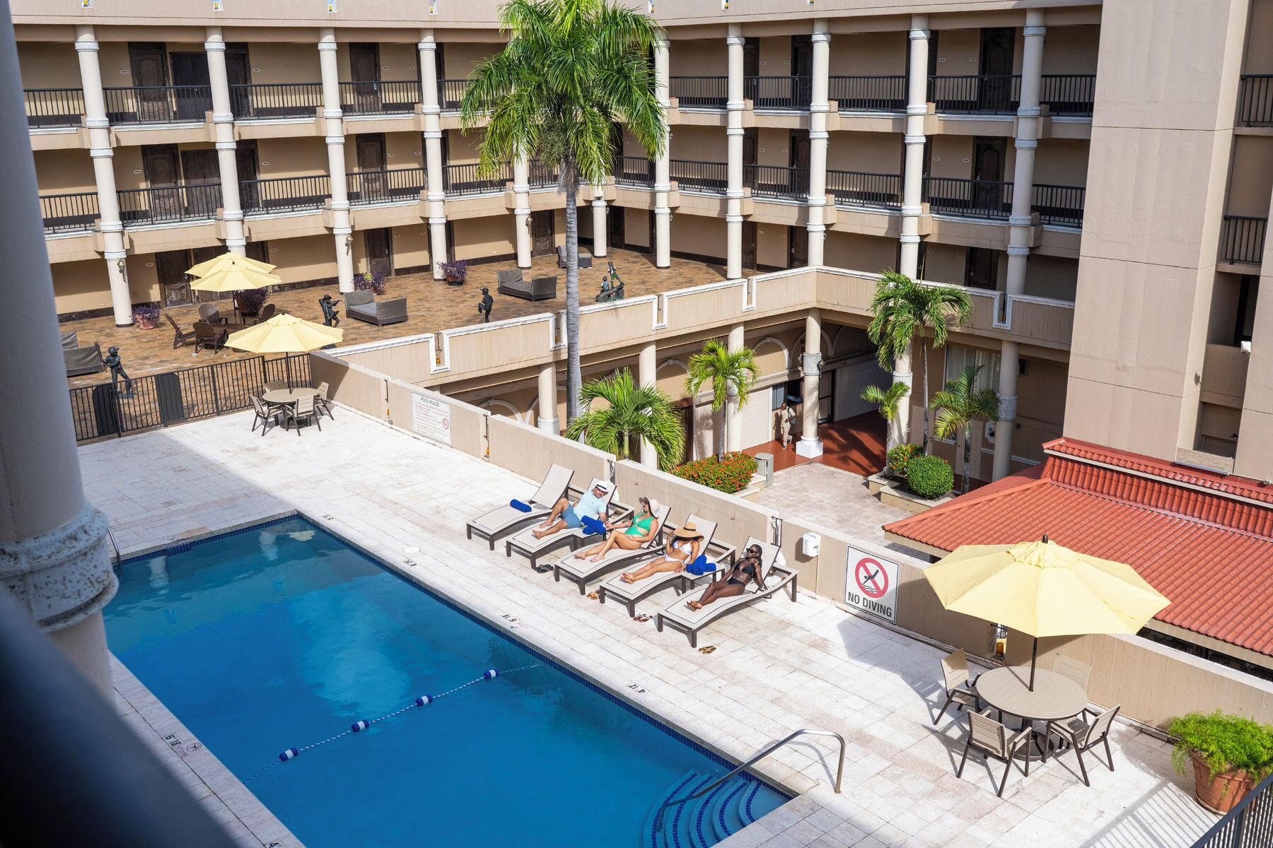 Un espace piscine d’hôtel comprenant de nombreux chaises longues et parasols, une piscine et un immeuble à plusieurs étages doté de balcons.