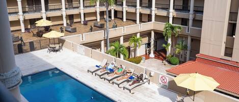 A hotel pool area with multiple lounge chairs and umbrellas, a swimming pool, and a multi-story building with balconies.